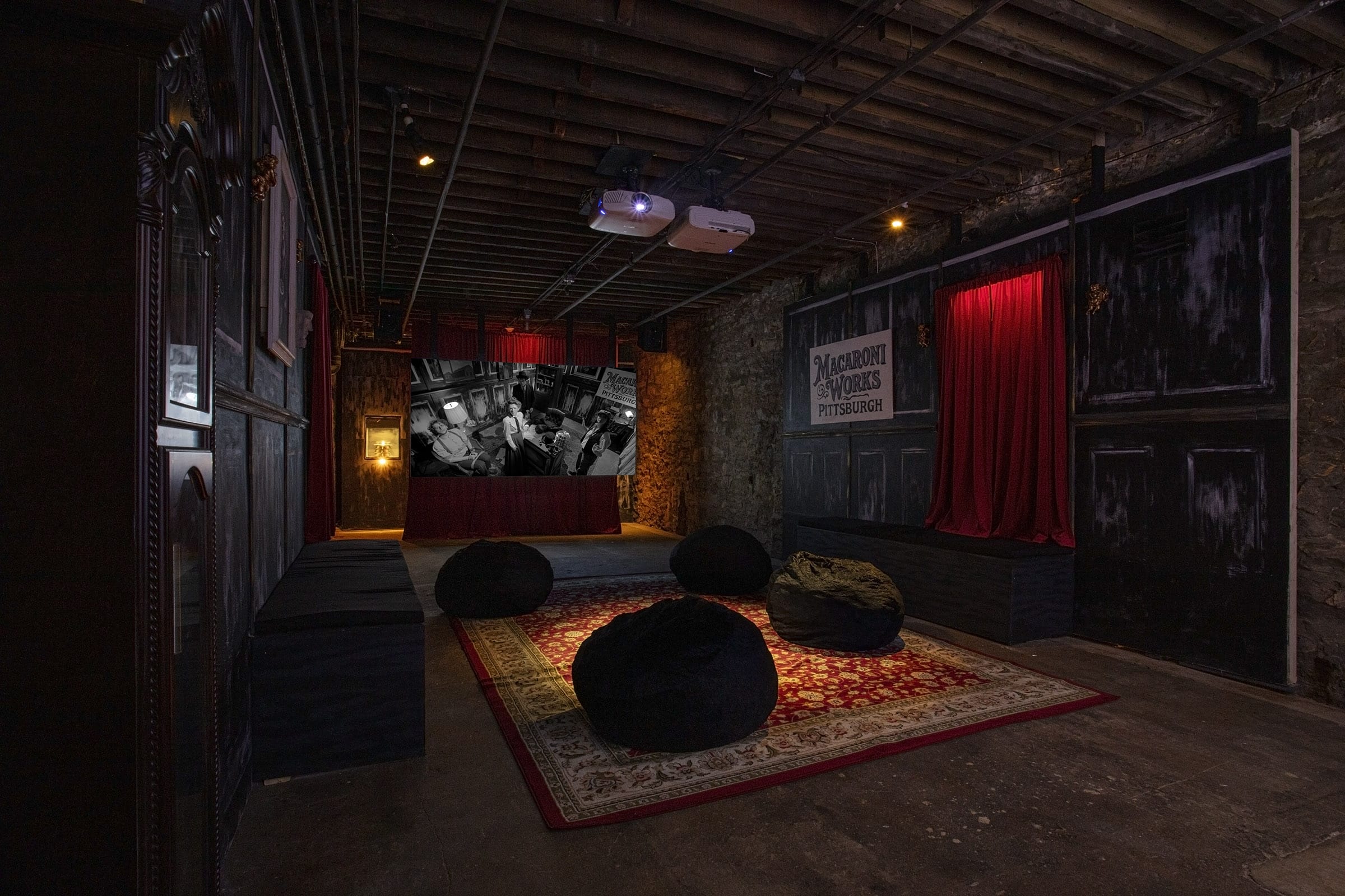 Image by Tom Little. An old-timey styled room with many red accents. It is dark. A black and white film plays on the back screen. There is a decorative rug and four black bean bags for people to sit on to watch the film.